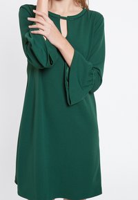 Grön, knälång klänning med rund halsringning, nyckelhålsdetalj och trekvarts ärmar med klockform. Slätt tyg med en svag lyster.