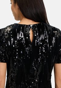 Czarna sukienka z wycięciem na plecach, pokryta błyszczącymi czarnymi i srebrnymi cekinami, o teksturowanym materiale, krótkich rękawach i dopasowanej sylwetce.