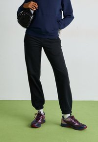 Juodos sportinės kelnės su elastiniais manžetais, derinamos su daugiaspalviais sportiniais batais su tinklinėmis viršutinėmis dalimis ir tekstūruota padu. Tamsiai mėlyna sportinė palaidinė ir maža juoda rankinė.