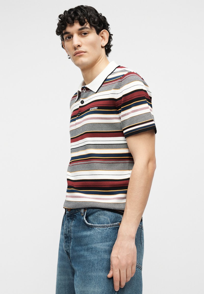 Young man wearing a short-sleeve striped polo shirt with white collar and blue jeans, standing against a plain light background.
