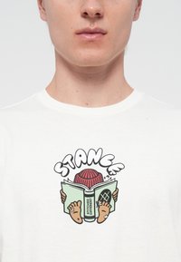 White cotton T-shirt featuring a colorful graphic print of a character reading a book, with bare feet, a red beanie, and the word "STANCE."