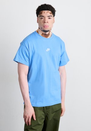 T-shirt en coton bleu clair avec un logo Nike blanc sur la poitrine, associé à un pantalon vert olive. Manches courtes et encolure ronde.