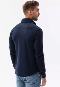 Maglietta navy a maniche lunghe con colletto, realizzata in un tessuto testurizzato, con polsini con bottoni e orlo curvo, mostrata da dietro.