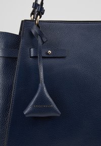 Laukkumalli on laivastonsininen nahkainen käsilaukku, jossa on teksturoitu pinta, kolmiomainen logotagi ja kontrastiväriset ompeleet. Kestäviä ketju- ja hihnakoristeita.