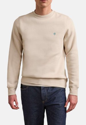 Homme portant un sweat-shirt beige à manches longues et col rond avec un petit logo d'arbre bleu sur le côté gauche de la poitrine, associé à un jean bleu foncé.