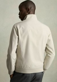 Jachetă bej deschis cu un guler înalt, textură netedă, buzunare laterale și manșete elastice. Viziunea din spate arată un design curat și minimalist.