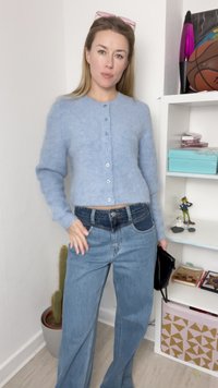 Cardigan azul claro e fofinho com mangas longas e botões, combinado com jeans de denim de cintura alta e pernas largas. Bolsa preta, decoração mínima ao fundo.