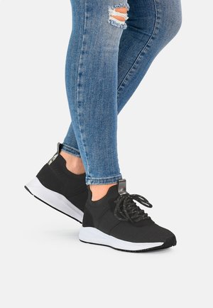 Ben iført slidte blå skinny jeans og sorte snørestøvler med hvide såler, stående mod en ensfarvet baggrund.