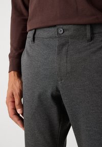 Hombre vestido con pantalones ajustados de color gris oscuro y una camisa de manga larga de color marrón oscuro, con la mano descansando relajada al lado.