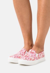 Slitna canvas-skor i vitt, med ett mönster av rosa och röda hjärtan. Gummisula med texturerat grepp, elastiska sidopaneler för lätt påtagning.