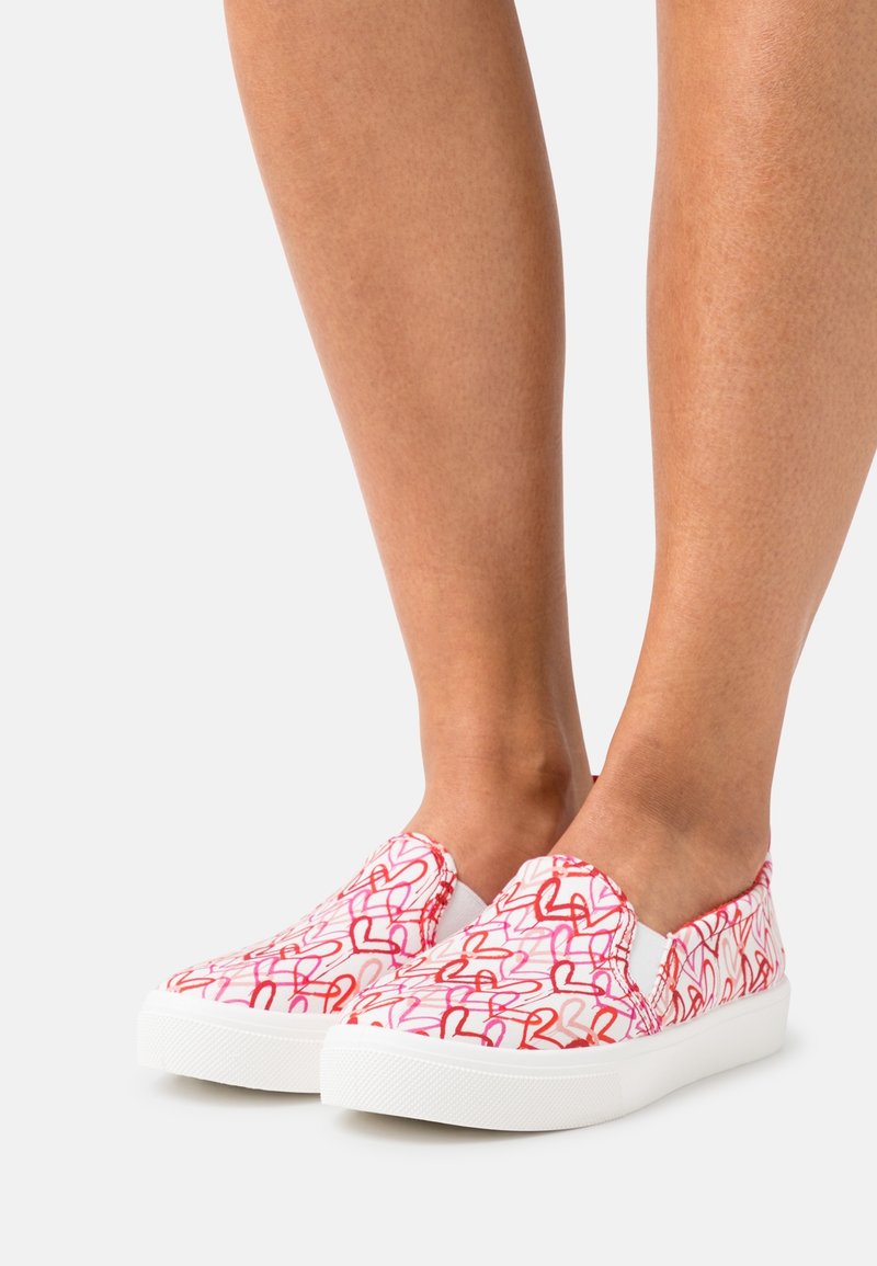 Slitna canvas-skor i vitt, med ett mönster av rosa och röda hjärtan. Gummisula med texturerat grepp, elastiska sidopaneler för lätt påtagning.