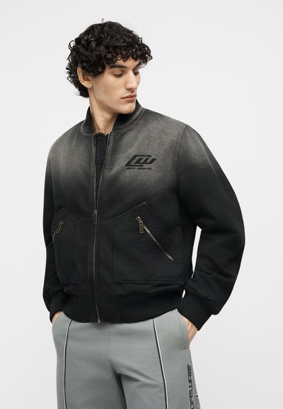 Άνδρας με σγουρά μαλλιά που φοράει μια μαύρη μπουφάν bomber με διαβαθμισμένο χρώμα, φερμουάρ σε τσέπες και γκρι παντελόνι με μαύρες ρίγες και κείμενο στο πλάι.