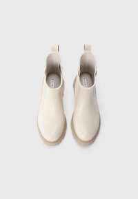 Bottines beiges en matériau brillant avec une semelle crantée, dotées d'un bout arrondi et d'une languette de traction de chaque côté.