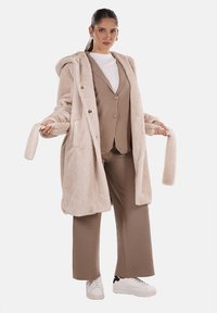 Cappotto in pelliccia sintetica beige con cappuccio, dotato di grandi bottoni, tasche e una consistenza morbida. Indossato sopra un completo marrone e una camicia bianca, abbinato a scarpe bianche.