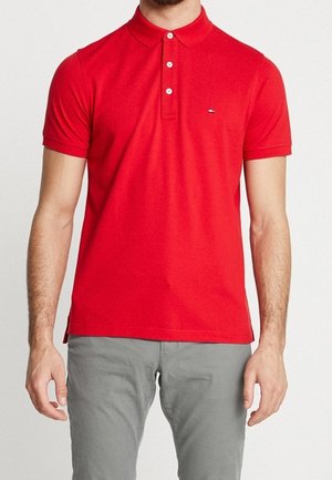 Mann trägt ein eng anliegendes rotes Poloshirt mit kurzen Ärmeln und drei weißen Knöpfen sowie graue Hose, steht vor einem schlichten weißen Hintergrund.