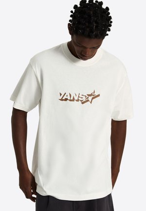 Man met kort krullend haar, draagt een wit Vans t-shirt met een bruin logo, kijkt naar beneden, staat tegen een effen lichte achtergrond.