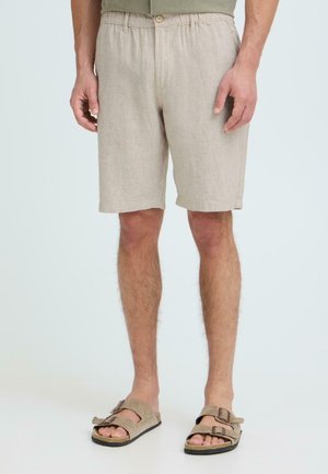 Mand iført beige knælange shorts og matchende beige sandaler med to remme og spænder, stående mod en ensfarvet baggrund.