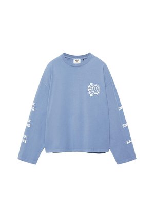 Lyseblå langærmet skjorte med hvidt rundt logo på brystet og hvid "KNWK" tekst gentaget på begge ærmer.