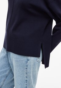 Marineblauer Strickpullover mit geripptem Saum, seitlichen Schlitzen und einem lockeren Design, kombiniert mit hellblauen Jeans.