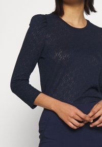 Tmavě modrý top s dlouhým rukávem a texturovaným vzorem, s nabíranými rameny a kulatým výstřihem. Měkká, lehká látka.