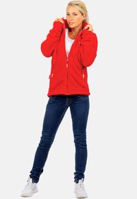 Roter Fleece-Zip-Hoodie mit Seitentaschen, mit einer strukturierten Oberfläche und gerippten Bündchen, kombiniert mit blauen Jeans und weißen Sneakers.