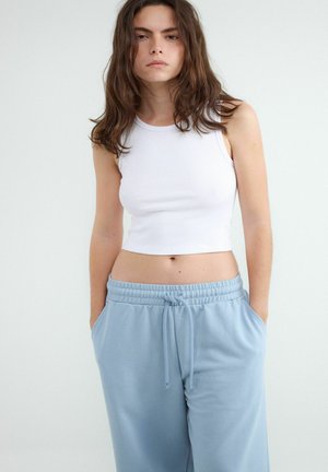 Młoda kobieta w białym bezrękawniku typu crop top i jasnoniebieskich spodniach dresowych z troczkiem, z rękami w kieszeniach na jednolitym tle.