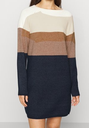 Vrouw die een gebreide jurk met lange mouwen draagt met horizontale kleurvlakken in wit, beige, bruin, lichtbruin en marineblauw.