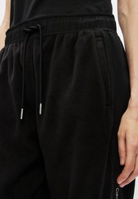 Czarne dresowe spodnie z polaru z elastycznym pasem i sznurkiem. Posiada boczne kieszenie i detal z logo wzdłuż nogawki. Miękka tekstura.