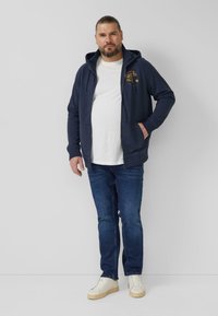 Marineblauwe hoodie met rits en zakken, een wit T-shirt eronder, blauwe spijkerbroek en witte sneakers. De hoodie heeft een logo op de borst.