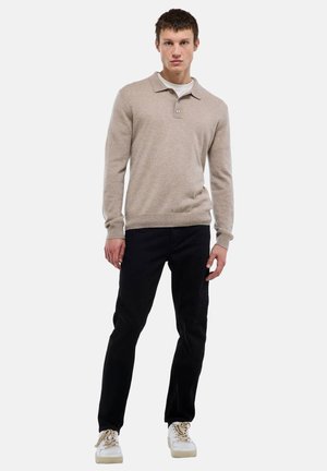 Jeune homme debout portant un pull polo beige à manches longues, pantalon noir et baskets blanches sur un fond clair uni.