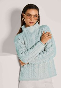 Frau trägt einen hellblauen Kabelstrick-Turtleneck-Pullover, goldene Creolen, braune Fliegerbrille und einen goldenen Ring, posiert mit verschränkten Armen.