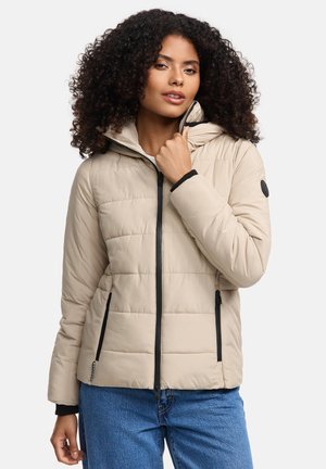 Vrouw met krullend haar in een beige gewatteerde jas met zwarte ritsdetails, die de kraag vasthoudt, gecombineerd met blauwe jeans.