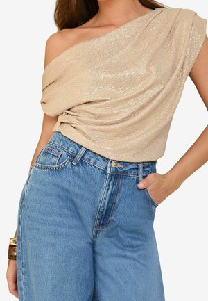 Kvinde iført en glitrende beigefarvet one-shoulder top, der er stoppet ned i højtaljede lyseblå jeans med den ene hånd i lommen.