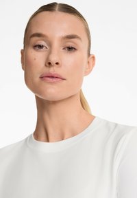Primo piano di una persona che indossa un top bianco in tessuto liscio, a girocollo. La pelle è chiara con trucco minimale e capelli lisci raccolti.