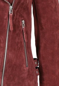 Bordeauxkleurig suède jack met zilveren ritsen aan de voorkant en zak, en twee verstelbare zijriemen met metalen oogjes.