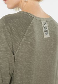 Olivegrünes, strukturiertes T-Shirt mit Rundhalsausschnitt, Label auf der Rückseite und Raglanärmeln, das ein dezentes meliertes Muster zeigt.