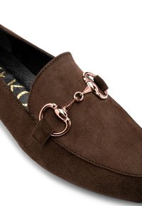 Mocassino in suede marrone con un dettaglio metallico, particolare hardware rotondo e cuciture tonali. Texture liscia con un design a suola piatta.