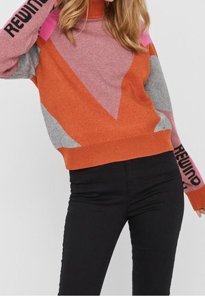 Femme portant un pull géométrique multicolore avec le texte "REWIND" sur les manches et un pantalon noir ajusté, debout devant un fond uni.