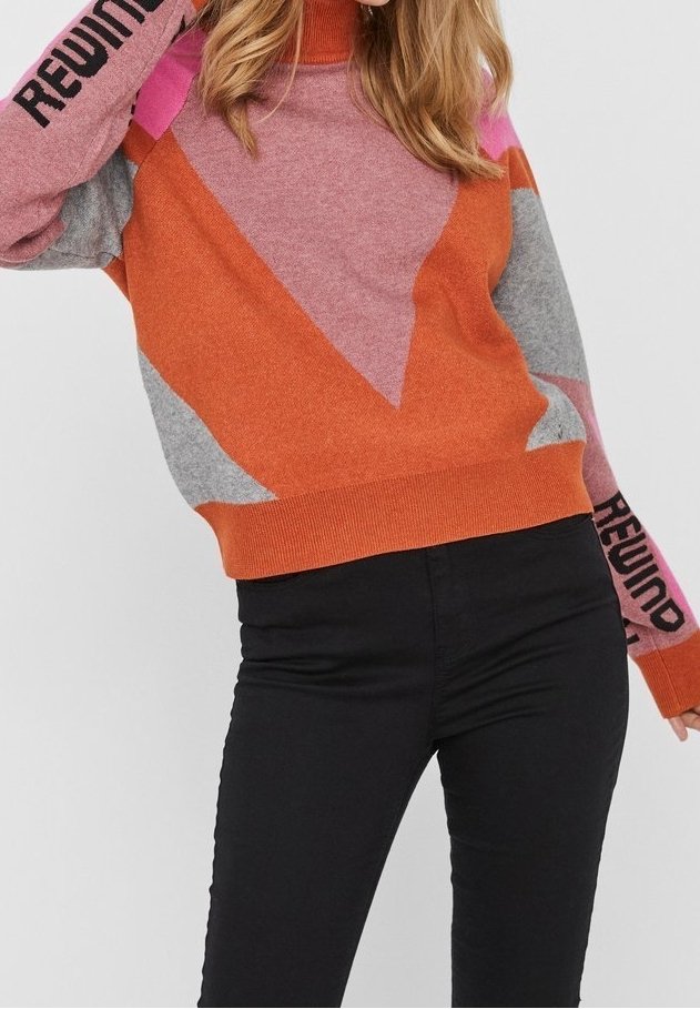 Femme portant un pull géométrique multicolore avec le texte "REWIND" sur les manches et un pantalon noir ajusté, debout devant un fond uni.