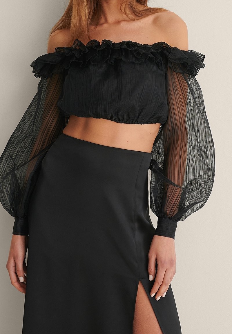 Femme portant un crop top noir épaules dénudées avec des manches ballon transparentes, associé à une jupe noire taille haute avec une fente sur la cuisse.