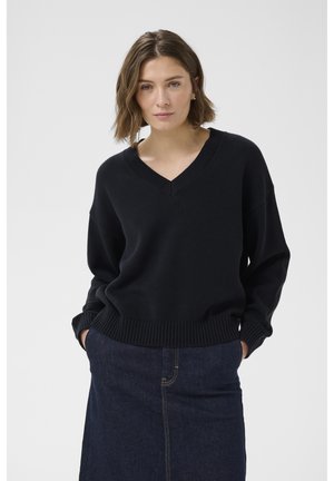 Rövid barna hajú nő fekete V-nyakú pulóverben és sötétkék farmer szoknyában, kezét zsebében tartva, fehér háttér előtt áll.