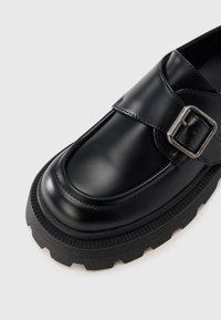 Svart lädersko med rundad tå, spännband och en klumpig gummisula med mönstrat grepp.