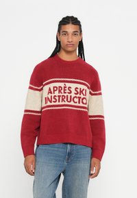 Persona con capelli lunghi e intrecciati che indossa un maglione rosso e beige con la scritta "APRÈS SKI INSTRUCTOR" e jeans blu, su uno sfondo bianco.