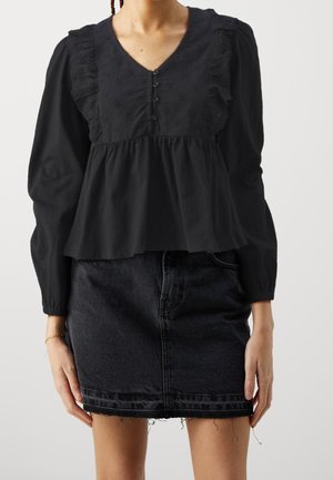 Vrouw draagt een zwarte blouse met ruches en knopen, gecombineerd met een zwarte denim minirok met rafelige zoom.