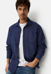 Young man with curly hair wearing a navy blue button-up shirt over a white t-shirt and blue jeans, standing against a white wall.