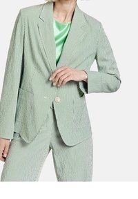 Blazer rayé vert et blanc avec une fermeture à un bouton, des revers crantés et deux poches avant. Associé à un top en satin vert.