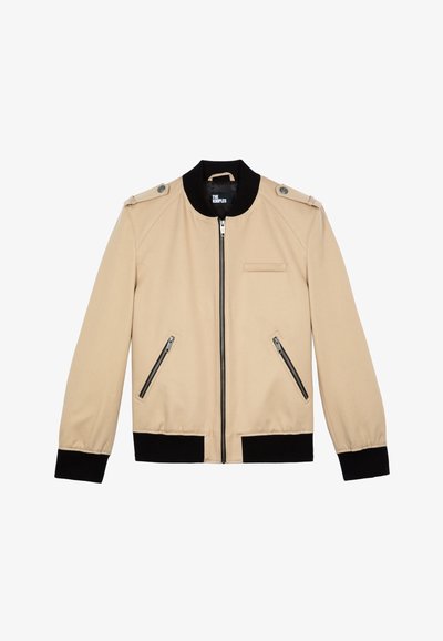 Beżowa kurtka bomberka z czarnymi ściągaczami przy mankietach i dolnej części; ma zamek błyskawiczny z przodu, dwie kieszenie na zamek i naramienniki.