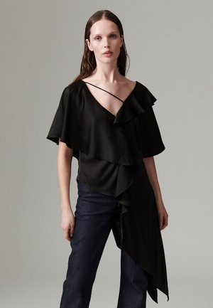 Femme aux longs cheveux bruns porte un top asymétrique noir avec des volants et un jean bleu foncé, debout devant un fond gris uni.