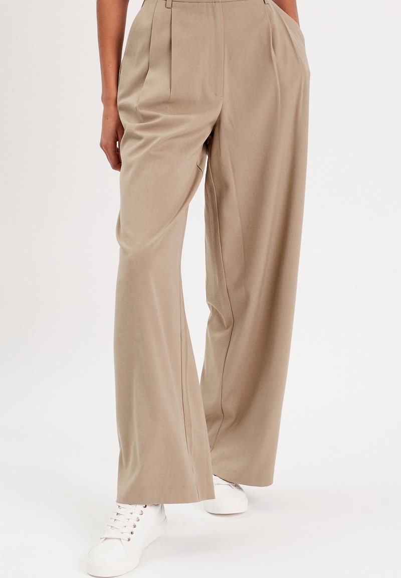 Pantalons beige à jambes larges en tissu lisse avec des plis. Dotés d'une taille haute et d'un design épuré et ajusté associés à des baskets blanches.