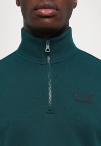 Detailný záber na osobu, ktorá má na sebe tmavozelený zipsový sveter s vysokým golierom a logom EA7 Emporio Armani na hrudi.